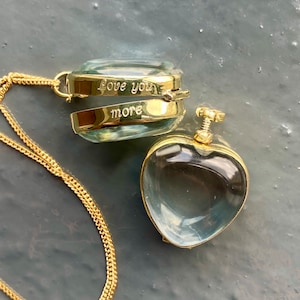 Op de afbeelding: Een gouden ketting met twee medaillon hangers. Het grotere medaillon is open en heeft de woorden "Love you more" gegraveerd aan de binnenkant. Het kleinere medaillon is hartvormig.