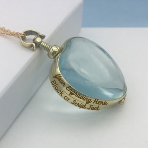 May include: A gold-colored heart-shaped pendant necklace with a light blue center. The pendant has the engraved text "Your Engraving Here Block or Script Font". The necklace is on a gold chain.