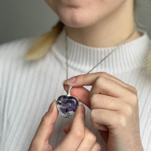 May include: A heart-shaped pendant necklace with a silver chain. The pendant is clear, filled with purple and white flowers, and has a silver bail. The necklace is being held up by a person.