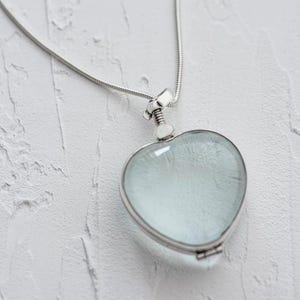 Op de afbeelding: Een zilveren ketting met een hartvormige hanger met een transparante glazen inzet. De hanger is open en de binnenkant is zichtbaar.