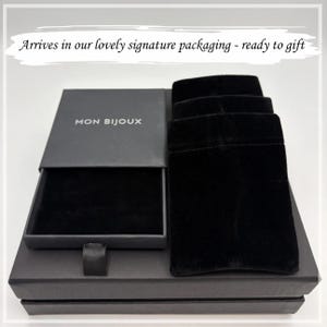 May include: A black rectangular jewelry box with a sliding drawer featuring the text MON BIJOUX in white lettering. Next to the box are three black velvet pouches arranged in a row. The set is presented on a black base with a white background.