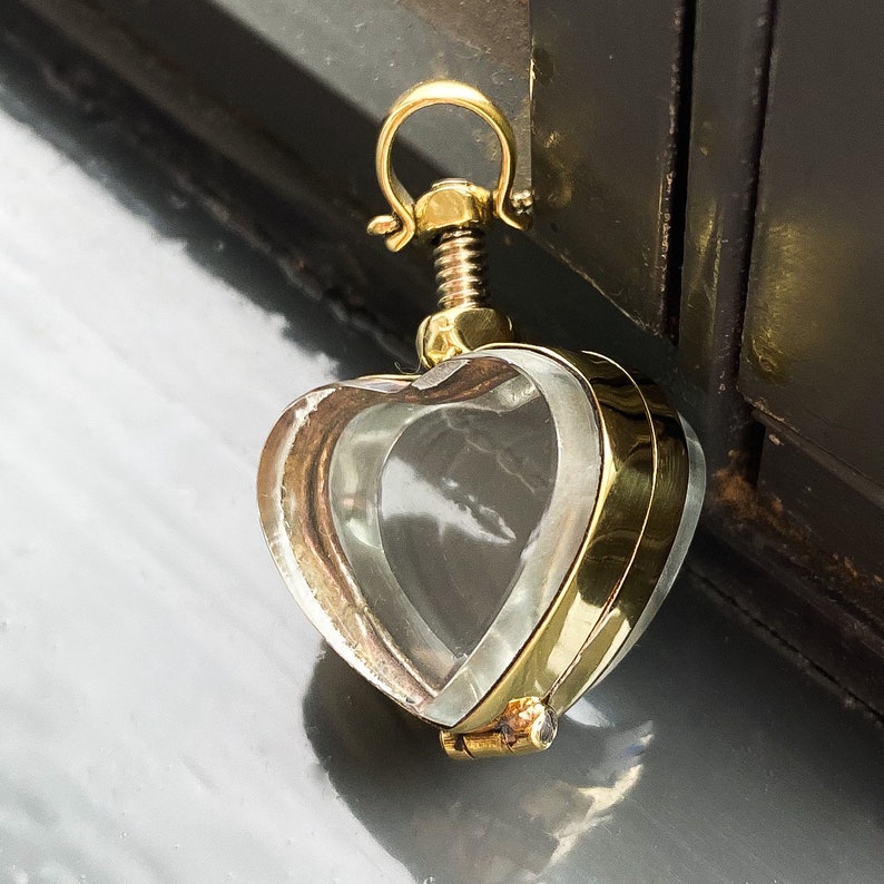 May include: A heart-shaped locket pendant featuring a clear glass or crystal center encased in a polished gold-tone metal frame. The top of the pendant includes a threaded screw mechanism and a circular bail for attachment to a chain or necklace.