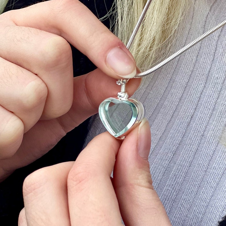 May include: A silver-toned heart-shaped locket pendant with a clear center, suspended from a thin silver-toned snake chain. The locket features a polished metallic frame surrounding a transparent heart-shaped insert, designed to hold small items.