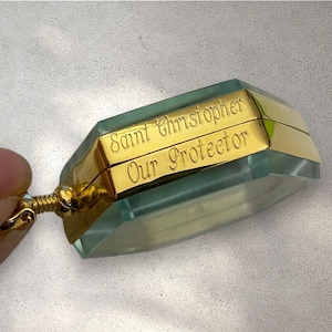 Puede incluir: Un colgante dorado con un dije de vidrio transparente de forma octogonal. El dije tiene las palabras "San Cristóbal Nuestro Protector" grabadas.