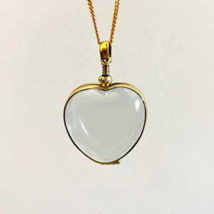 May include: A heart-shaped pendant featuring a clear, smooth glass or crystal center encased in a polished gold-tone metal frame. The pendant hangs from a delicate gold-tone chain and includes a small, threaded screw-top mechanism at the top.