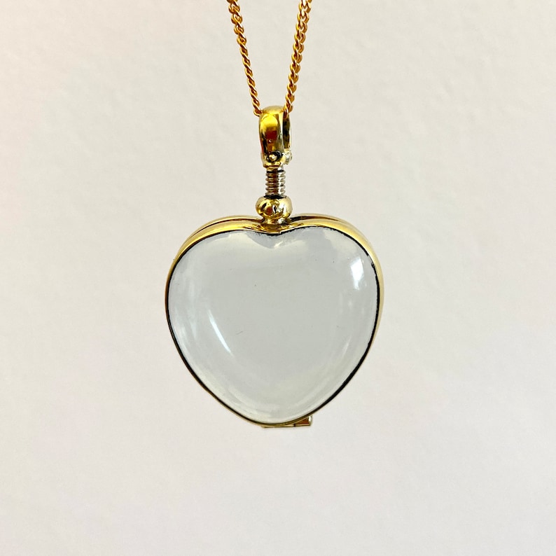 May include: A heart-shaped pendant featuring a clear, smooth glass or crystal center encased in a polished gold-tone metal frame. The pendant hangs from a delicate gold-tone chain and includes a small, threaded screw-top mechanism at the top.