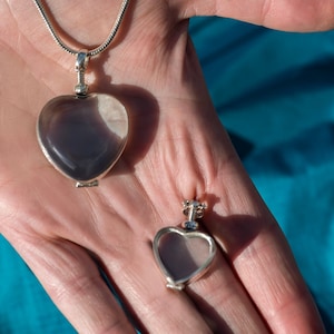 May include: A silver-toned heart-shaped locket pendant with a smooth, translucent grey stone inlay, attached to a silver-toned chain. A smaller, matching heart-shaped locket frame without the stone inlay is positioned separately below the larger pendant.