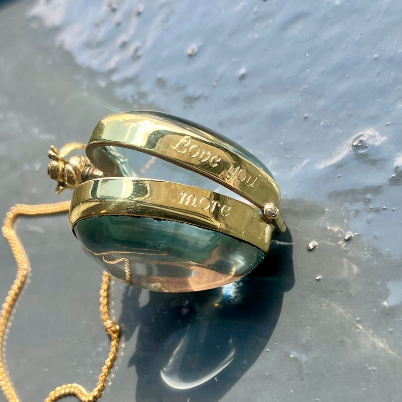 May include: A gold locket pendant with a clear, rounded glass orb. The locket is engraved with the words "Love you more". A delicate gold chain is attached to the pendant.