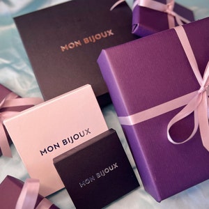 May include: A selection of jewellery boxes in shades of black, pink, and purple. Each box is branded with the text "MON BIJOUX" and some are adorned with pink ribbons. The boxes are presented on a light blue surface.