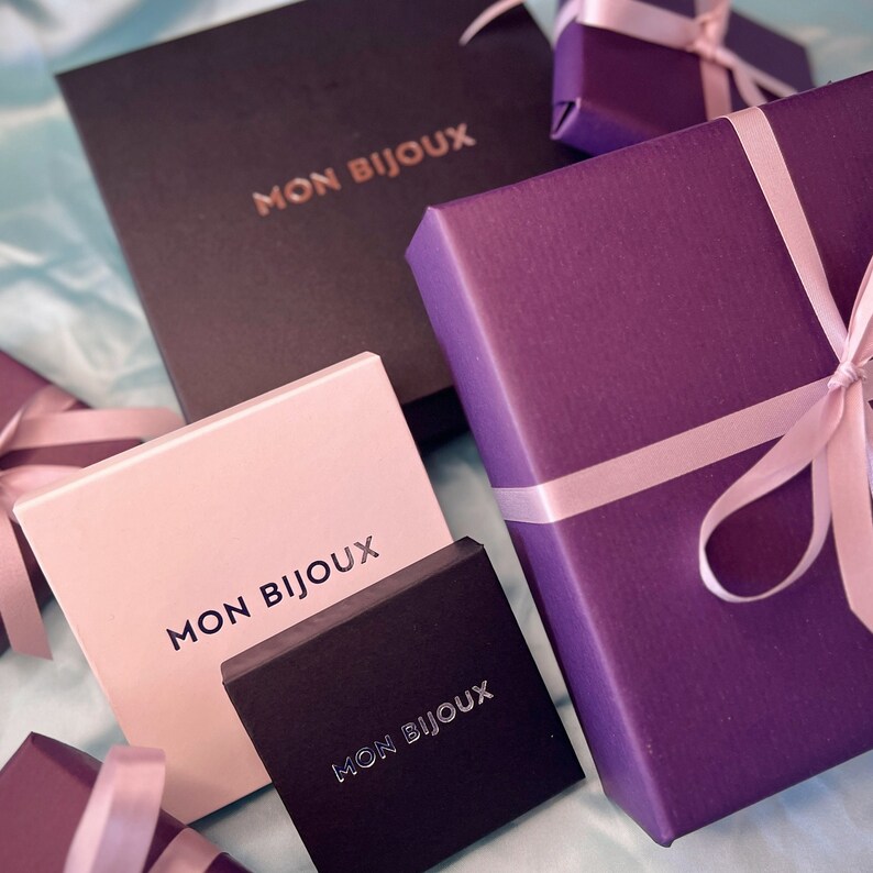 May include: A selection of jewellery boxes in shades of black, pink, and purple. Each box is branded with the text "MON BIJOUX" and some are adorned with pink ribbons. The boxes are presented on a light blue surface.