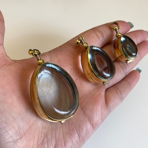 May include: Three gold oval-shaped locket pendants with clear glass windows. The lockets are of different sizes and are hanging from a hand.