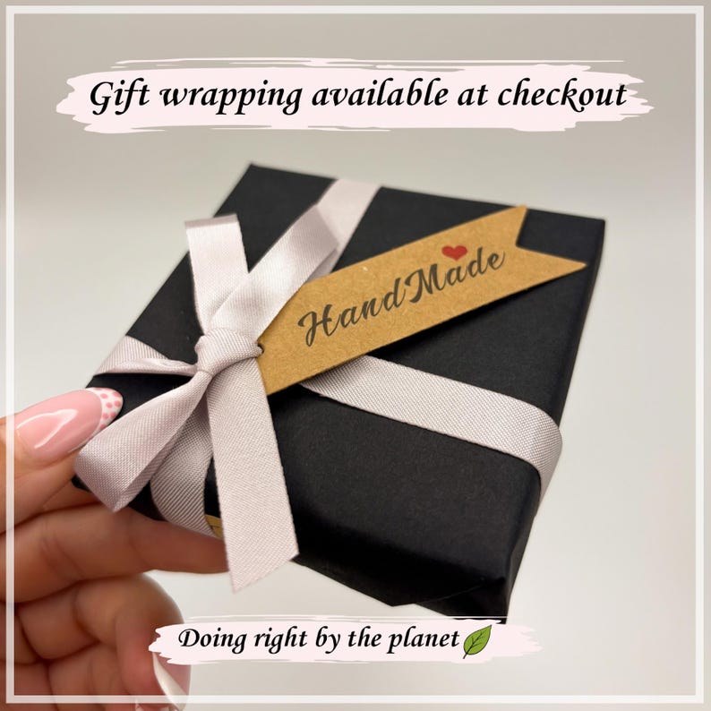 May include: A black gift box tied with a light grey ribbon and bow. A small brown tag with "Hand Made" and a red heart is attached. The box is wrapped in black paper.