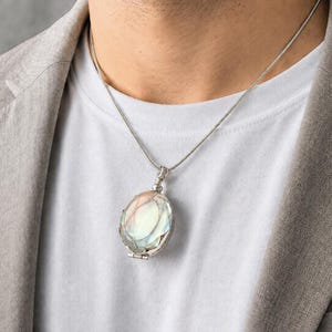 May include: A silver-toned locket pendant featuring a smooth, iridescent oval stone with a pearlescent sheen, suspended from a thin, matching silver-toned snake chain necklace that rests against a plain white t-shirt and a light gray blazer.