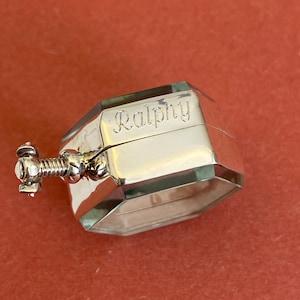 Op de afbeelding: Een zilveren achthoekige doos met een scharnierend deksel. Het deksel is gegraveerd met de naam "Ralphy". De doos heeft een klein, zilveren schroefslot.