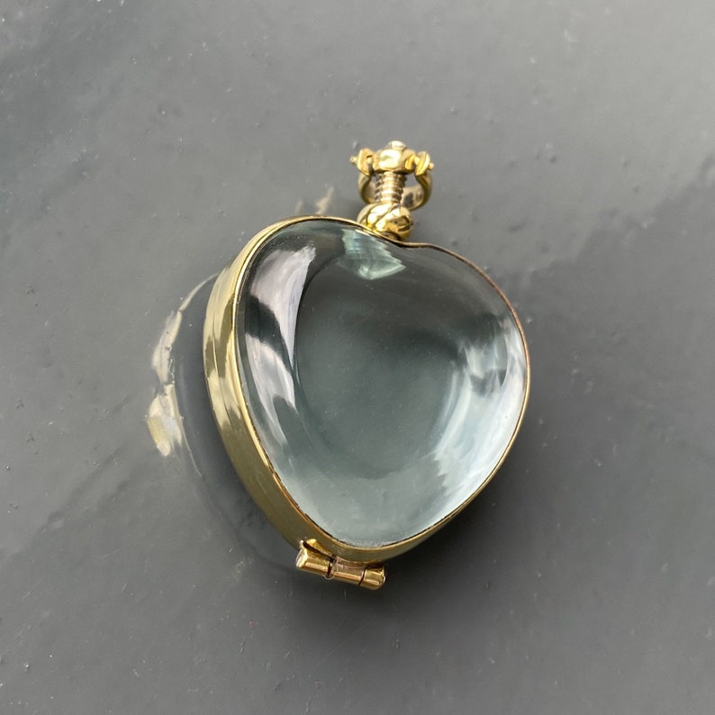 May include: A heart-shaped locket featuring a clear, smooth glass or crystal front encased in a polished gold-tone metal frame. The piece includes a small, hinged metal bail at the top for attachment, showcasing a reflective surface and metallic edges.