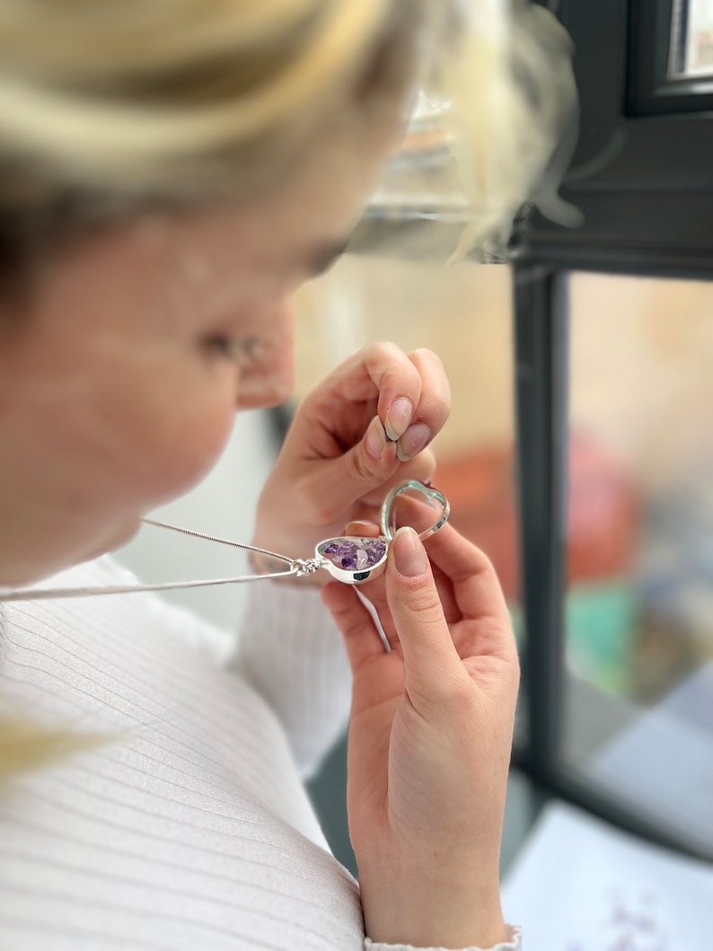 May include: A silver heart-shaped locket necklace filled with small purple gemstones. The locket is open, revealing the gemstones inside. The necklace is held by a person wearing a white shirt.