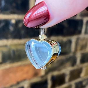 May include: A heart-shaped pendant with a clear, light blue interior and gold-colored trim. The pendant has a screw-top closure and is suspended from a gold-colored metal piece. The background is a blurred brick wall.