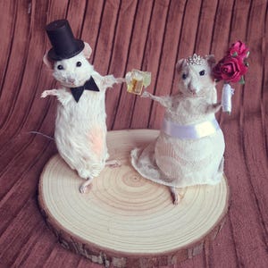 May include: A whimsical wedding cake topper featuring two white mice. The mouse on the left wears a black top hat and bow tie, holding a miniature drink. The mouse on the right wears a bridal gown, tiara, and holds a bouquet of pink roses. They stand on a wooden slice.
