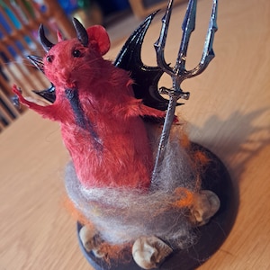 May include: A red, furry devil rat figurine with black wings and horns, holding a silver trident. The figurine is sitting on a black base with a white and orange fuzzy material around it.