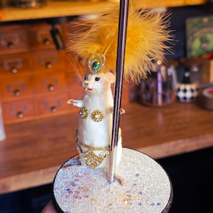 May include: A white taxidermied mouse wearing a gold sequined costume and a yellow feather headdress, posed on a silver glitter platform with a silver pole.