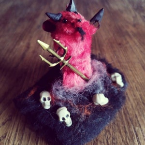 May include: A red plush devil figurine holding a gold trident, sitting on a black and orange fabric base with white skull accents.