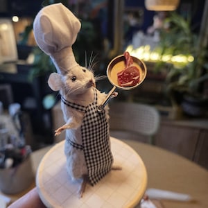 May include: A taxidermied mouse in a chef's outfit, including a white hat and a black and white checkered apron. The mouse holds a small pan with a piece of cooked meat. It stands on a round wooden base.