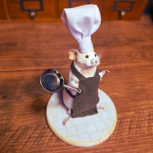 May include: A taxidermied white mouse figurine dressed as a chef, wearing a white chef's hat and a brown apron. The mouse holds a small silver ladle. It stands on a round, tiled base.