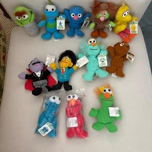May include: A collection of twelve Sesame Street plush toys including characters like Elmo, Cookie Monster, Big Bird, and Count von Count. Several figures feature attached paper tags with the Sesame Street logo and character names.