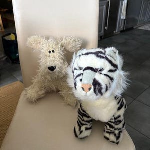May include: Two plush toys sit on a chair. One is a cream-colored dog with floppy ears and a black nose. The other is a white tiger with black stripes, a pink nose, and blue eyes. The toys are soft and cuddly.