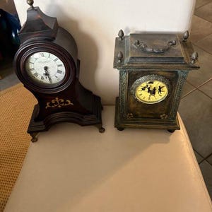 May include: Two antique clocks. The first is a dark wood mantel clock with a round face and ornate details. The second is a square clock with a bronze finish and a decorative handle on top. Both clocks have visible clock faces.