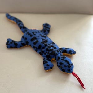 May include: A blue and black spotted plush lizard toy with a red tongue. The lizard is lying on its belly with its legs outstretched.