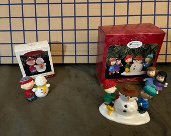 May include: Two Peanuts Christmas ornaments, one featuring Charlie Brown and Snoopy building a snowman, the other featuring the Peanuts gang building a snowman. The ornaments are packaged in their original boxes.
