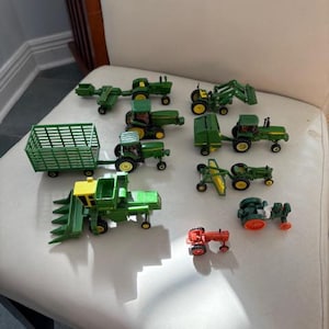 May include: A collection of toy farm vehicles, including tractors, harvesters, and trailers. The toys are primarily green and yellow, with a small red tractor. The toys are arranged on a white surface.