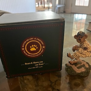 May include: A Boyds Bears & Friends figurine of a brown bear wearing a baseball uniform and a blue cap with a white 'B' on it. The bear is holding a baseball and is in a pitching position. The figurine is on a base with green grass and brown dirt. The figurine is in a box with the text "Boyds Bears & Friends The Bearstone Collection... Bears & Hares you can Trust... est. 1979" on it.