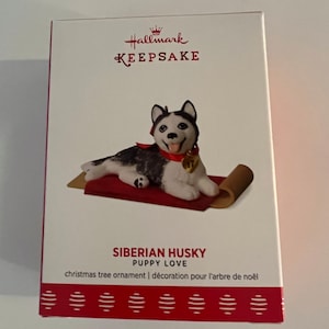 K&ouml;nnte beinhalten: Hallmark Keepsake Weihnachtsbaumschmuck mit einem sibirischen Husky-Welpen, der auf einem roten und braunen Teppich liegt. Der Anh&auml;nger tr&auml;gt den Titel "Siberian Husky Puppy Love" und den Text "christmas tree ornament | d&eacute;coration pour l'arbre de no&euml;l".
