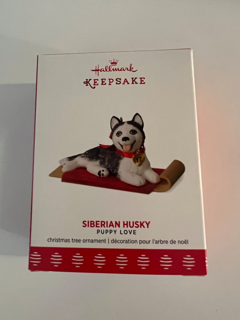 K&ouml;nnte beinhalten: Hallmark Keepsake Weihnachtsbaumschmuck mit einem sibirischen Husky-Welpen, der auf einem roten und braunen Teppich liegt. Der Anh&auml;nger tr&auml;gt den Titel "Siberian Husky Puppy Love" und den Text "christmas tree ornament | d&eacute;coration pour l'arbre de no&euml;l".