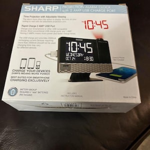 May include: A Sharp projection alarm clock with a 2 AMP USB charge port. The clock displays the time in red digits, with the time 10:45 visible. The clock also displays the date and time in a digital format. The box includes the instruction manual.