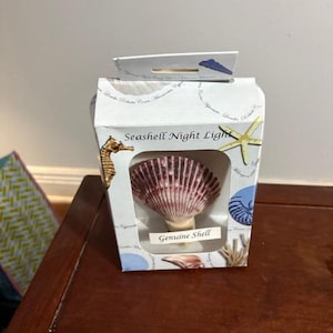 May include: A seashell night light in a white box with a clear window. The shell is a deep maroon color with white ridges. The box has illustrations of a seahorse, starfish, and other sea life.