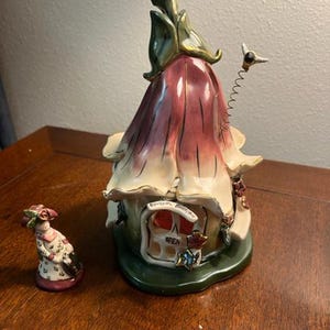 May include: A whimsical ceramic fairy house with a flower-like design, featuring a burgundy and cream color scheme. The house has an open door and the words "Boutique" and "Open" are visible. A small figurine of a fairy is next to the house.