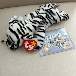 May include: White and black striped stuffed tiger toy with a red and white Ty Beanie Baby tag. The tag says "Beanie Baby" and has a star on it. The tiger is lying on its side with its head resting on its paws. The tiger is wearing a white ribbon around its neck. The tiger is on a light brown surface. The tag is attached to the tiger's right paw. There is a clear plastic card with a picture of the tiger on it. The card says "Blizzard the Tiger" and "Beanie Babies" on it. The card also says "Series 1 4163 1998" on it.