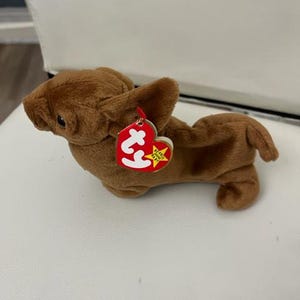 May include: Brown plush dog toy with a red "Ty" tag attached. The tag has a yellow star and the words "Beanie Babies" printed on it.