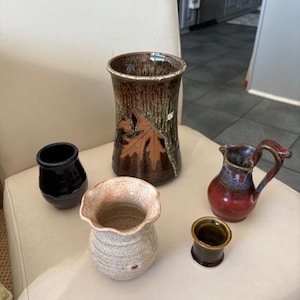 May include: A collection of handcrafted ceramic vases and pitchers. The pieces vary in size, shape, and color, including a tall brown vase with a leaf design, a small black vase, a red pitcher, and a small beige vase.