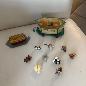 May include: A wooden ark-shaped toy with a green base and red wheels, accompanied by miniature animal figurines. The ark has a light brown wooden exterior and a green roof. The animals include penguins, giraffes, and lions.
