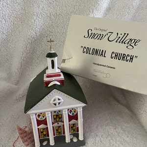 May include: A hand-painted ceramic church figurine from the Snow Village collection. The church is red and white with a green roof and a gold cross on top. The text "Colonial Church" is printed on the box.