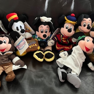 May include: A collection of nine plush Mickey Mouse toys, each dressed in a different costume. The costumes include a wizard, a conductor, a Hawaiian shirt, a British guard, a Roman toga, a cowboy, a pilot, a graduation gown, and a chef. The toys are all sitting on a brown leather couch.