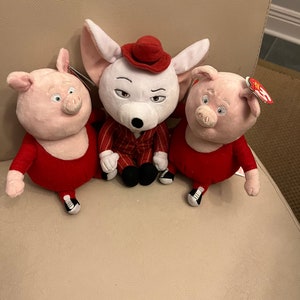 May include: Three plush toys: a white mouse wearing a red hat and a red and white striped suit, and two pink pigs wearing red outfits. The toys are sitting on a light brown surface.