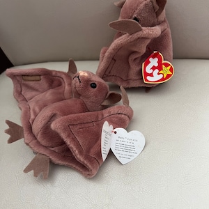 May include: Two brown plush bat Beanie Babies. The larger bat is lying on its back with its wings spread out. The smaller bat is sitting upright with its wings folded. Both bats have a red Beanie Baby tag attached.