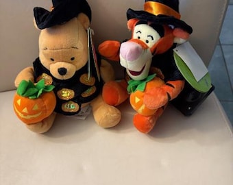 May include: Two plush Halloween-themed toys. One is a yellow bear wearing a black witch hat and vest, holding an orange pumpkin. The other is an orange and black tiger wearing a black witch hat and holding an orange pumpkin.