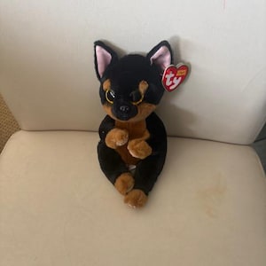May include: A plush Chihuahua dog toy with black and brown fur, large gold eyes, and a red heart-shaped tag. The toy is sitting upright on a white surface.
