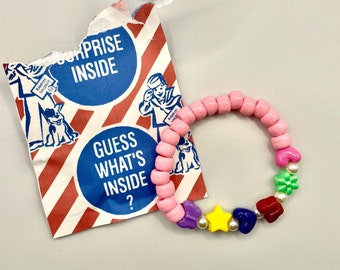 Puede incluir: Un brazalete de cuentas rosadas con abalorios coloridos, incluyendo corazones, estrellas y un trébol de cuatro hojas. El brazalete está al lado de un trozo de papel rasgado con el texto "Surprise Inside" y "Guess What's Inside?"